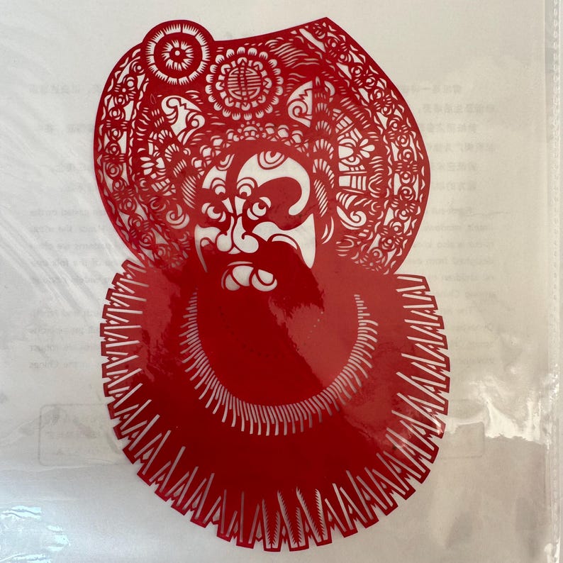 May include: A red paper cut-out depicting a traditional Chinese opera figure. The figure has a detailed headdress and beard, with intricate patterns and designs. The cut-out is likely used for decoration or as a cultural craft.