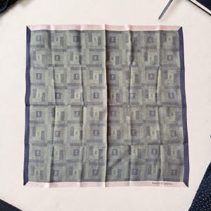 May include: A square pocket square featuring a repeating geometric pattern in shades of grey and dark blue. The fabric has a light pink border on the top and bottom edges, with the text Roberta di Camerino printed in the bottom right corner.
