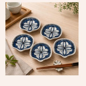 Peut inclure: Un ensemble de cinq petits plats en céramique en forme de coquillage, dotés d'une base blanche avec un motif floral bleu peint à la main. Chaque plat présente un bord festonné et un motif central en croix. Des baguettes reposent sur un support.