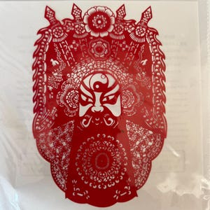 May include: A red paper cut-out depicting a traditional Chinese opera mask. The mask features a white face with black and red details, a long white beard, and elaborate headwear. The intricate design is visible against a white background.