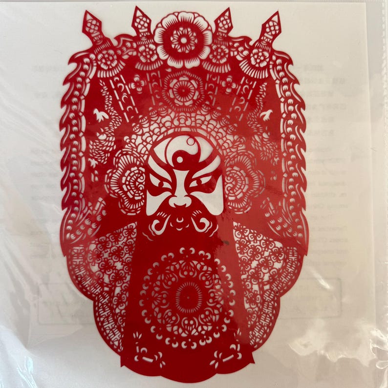 May include: A red paper cut-out depicting a traditional Chinese opera mask. The mask features a white face with black and red details, a long white beard, and elaborate headwear. The intricate design is visible against a white background.