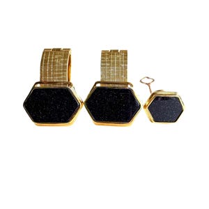 May include: Two gold-toned metal cuff links with textured bands and hexagonal faces featuring dark blue, glittery stone inlays, accompanied by a matching smaller hexagonal lapel pin with a thin gold-toned metal post and a small circular backing.