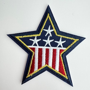 May include: Embroidered star patch featuring a navy blue border with a yellow outline. The patch has three white stars at the top and red and white stripes at the bottom, resembling a flag design. The patch is likely for clothing or accessories.