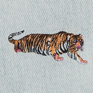 May include: Embroidered patch of a tiger in shades of orange, black, and white, with pink accents on the paws and face. The tiger is depicted in a running pose, with its tail curved. The patch is on a light blue denim background.