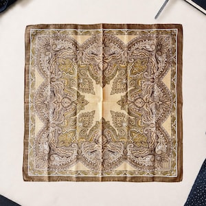 May include: A square pocket square featuring a symmetrical paisley pattern in shades of brown, olive green, and cream. The design includes intricate swirling motifs and floral-inspired shapes arranged in a border, finished with a thin white line.