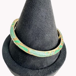 May include: A gold-toned hinged bangle bracelet featuring a decorative pattern of light green enamel inlay. The metal surface has a polished finish, and the green enamel sections are distributed evenly along the circumference of the circular band.
