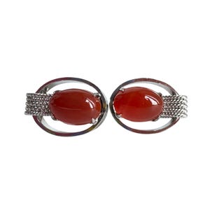 May include: A pair of oval-shaped cufflinks featuring polished, reddish-orange stones set in silver-toned metal. The design includes a woven texture on the sides, adding detail to the jewelry.