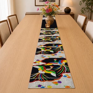 Peut inclure: Un long chemin de table rectangulaire présentant un motif répétitif d'oiseaux phénix colorés sur fond noir, alternant avec des sections de tissu gris clair décorées de petits motifs floraux vibrants orange, jaunes et violets.