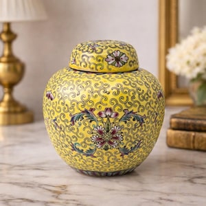 May include: A yellow ceramic ginger jar with a lid, adorned with a floral and scroll pattern. The jar features a central floral design with red, purple, and green accents. The background includes a gold lamp and a framed mirror.