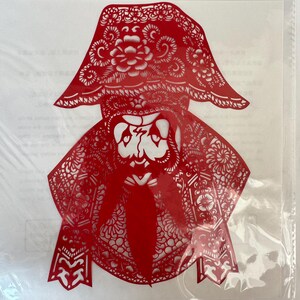 May include: A red paper cut-out of a figure with a hat and long beard. The intricate design features floral and geometric patterns, creating a detailed silhouette. The figure appears to be wearing a hat with a floral design.