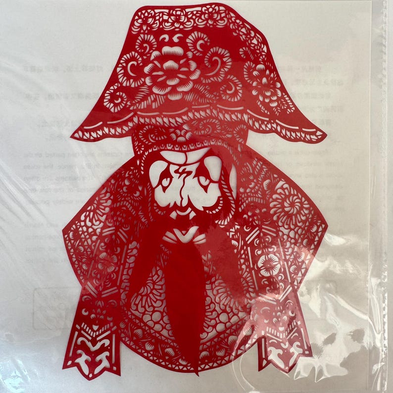 May include: A red paper cut-out of a figure with a hat and long beard. The intricate design features floral and geometric patterns, creating a detailed silhouette. The figure appears to be wearing a hat with a floral design.
