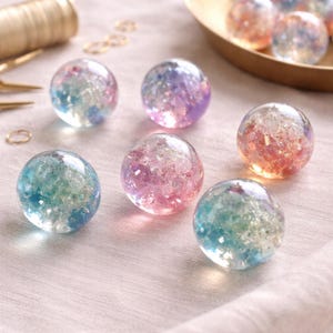 May include: A collection of clear, spherical orbs filled with colorful glitter. The orbs display hues of blue, pink, purple, and orange. Some orbs are scattered on a light pink surface, while others are in a gold-colored dish.