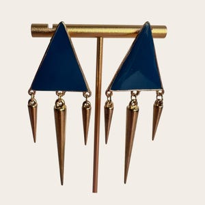 May include: A pair of geometric earrings. Each earring features a navy blue triangle with a gold-colored border and three gold-colored cone-shaped dangles. The earrings are displayed on a gold-colored earring stand.