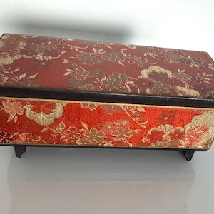 May include: Rectangular red jewelry box with a floral pattern in gold. The box has a dark wooden frame and sits on small black feet. The lid and sides are covered in a patterned fabric.