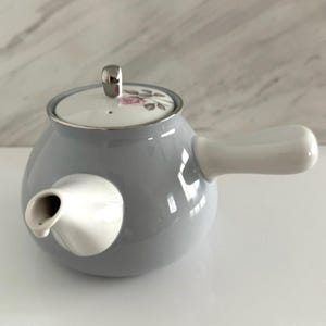 May include: A gray ceramic teapot with a white handle and spout. The lid is white with a floral design and a silver knob. The teapot has a rounded body and a smooth, glossy finish. A classic design for serving tea.