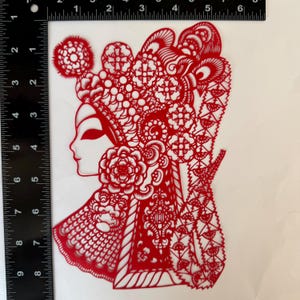 May include: A red paper cut-out depicting a woman's profile, adorned with an elaborate headdress and floral details. The intricate design showcases traditional Chinese art, with delicate patterns and shapes. The artwork is approximately 6 inches tall.