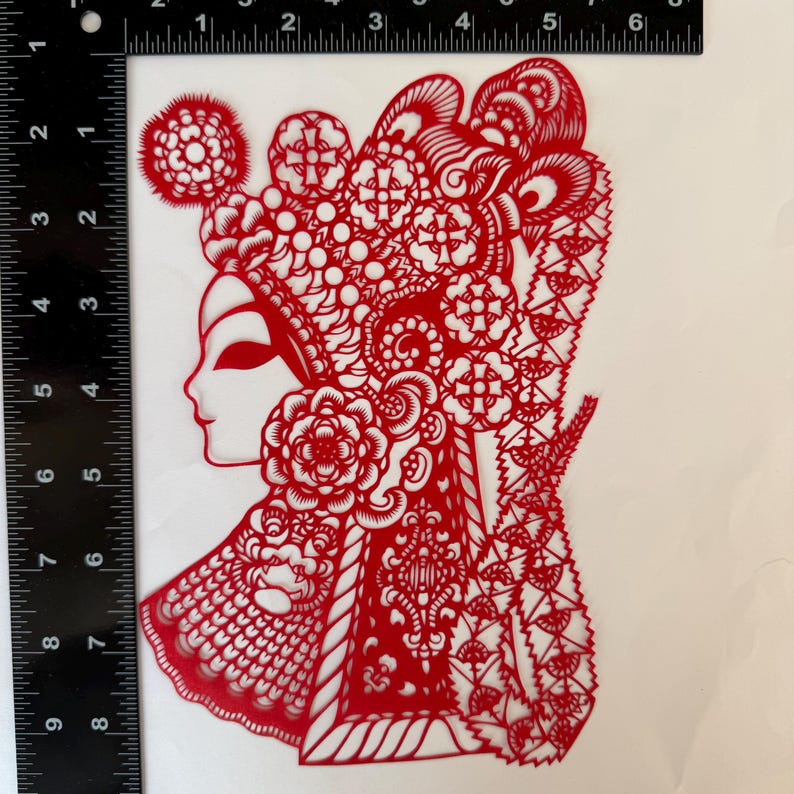 May include: A red paper cut-out depicting a woman's profile, adorned with an elaborate headdress and floral details. The intricate design showcases traditional Chinese art, with delicate patterns and shapes. The artwork is approximately 6 inches tall.