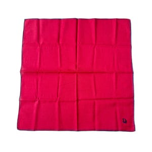 May include: A bright red handkerchief with a dark blue border and a small, dark blue logo in the corner. The fabric is folded into a square shape, with visible creases. The handkerchief is likely made of a soft, lightweight material.