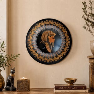 May include: A black decorative plate with an Egyptian pharaoh design. The plate features a gold and orange pharaoh profile in the centre, surrounded by gold and silver accents. The plate is displayed on a wooden surface with other Egyptian-themed decor.