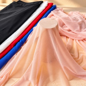 Könnte beinhalten: Eine Sammlung gefalteter Stoffstücke in Schwarz, Weiß, Rot und Blau, mit einem transparenten, hellpfirsichfarbenen Stoff, der über einen zylindrischen Holzständer im Vordergrund drapiert ist und weiche Falten sowie eine durchscheinende Optik erzeugt.