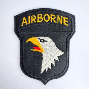 May include: A black patch with the word "AIRBORNE" in yellow arched across the top. Below the text is a white eagle head with a yellow beak and red tongue. The patch has a black border.