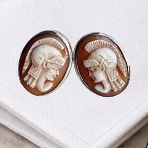 May include: A pair of oval-shaped cameo earrings featuring white carved profiles of figures wearing plumed helmets set against a reddish-brown background, encased in a polished silver-toned metal frame with a smooth, rounded edge finish.