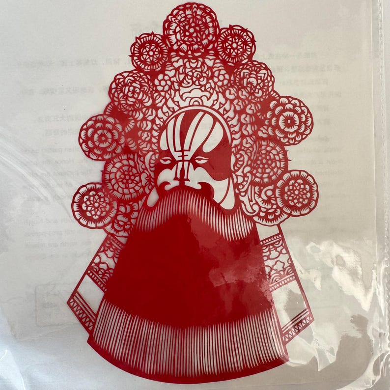 May include: Red paper cut-out of a traditional Chinese opera mask. The mask features a white face with red accents, a long white beard, and an elaborate headdress with floral and geometric patterns. The cut-out is in a clear plastic sleeve.