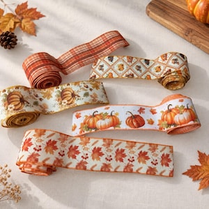 May include: Five rolls of decorative ribbon featuring autumn themes. One roll has an orange and brown plaid pattern. Others display white backgrounds with printed orange pumpkins, fall leaves, berries, and diamond-shaped pumpkin motifs.