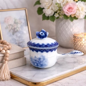 May include: A white and blue porcelain tea warmer with a handle and a lid topped with a blue rose. The tea warmer has a blue floral design and sits on a marble tray with a gold edge. A framed picture and flowers are in the background.