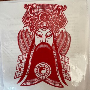 May include: A red paper cut-out of a traditional Chinese opera figure. The figure has a long beard, elaborate headdress, and a yin-yang symbol. The intricate design is visible against a white background.