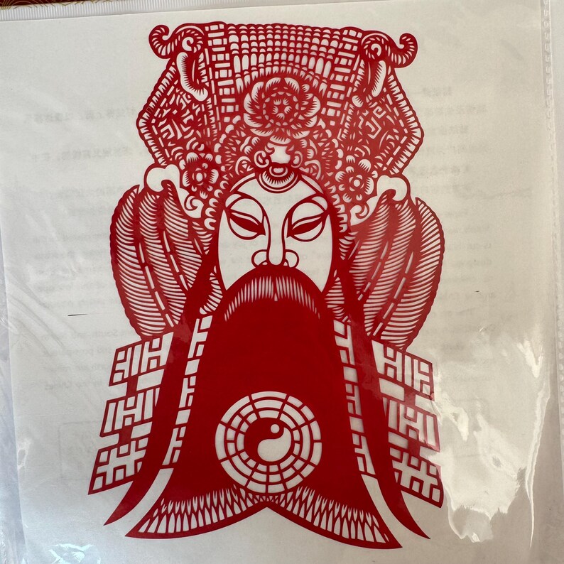 May include: A red paper cut-out of a traditional Chinese opera figure. The figure has a long beard, elaborate headdress, and a yin-yang symbol. The intricate design is visible against a white background.