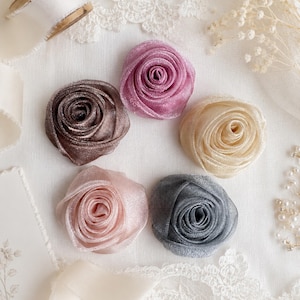 May include: Five fabric rose embellishments arranged in a circle. The flowers are made of a shimmering, translucent material in shades of mauve, dark brown, cream, pale pink, and slate gray, each featuring tightly coiled petals in a spiral design.