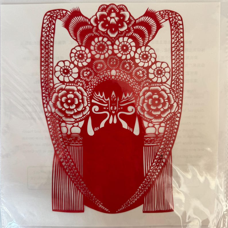 May include: A red paper cut-out of a traditional Chinese opera mask. The intricate design features floral and geometric patterns, with a central mask shape and decorative elements. The artwork is displayed on a white background.