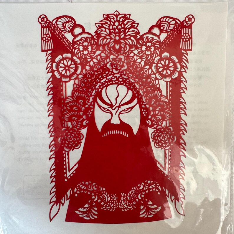 May include: A red paper cut-out of a Chinese opera mask. The mask features a detailed design with floral and geometric patterns, a long beard, and a stern expression. The cut-out is enclosed in a clear plastic sleeve.