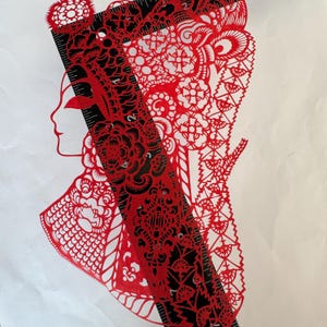 May include: A red paper cut silhouette of a woman's profile, adorned with intricate floral and geometric patterns. A black ruler is placed diagonally across the design. The artwork is set against a white background, showcasing the detailed cut-out design.