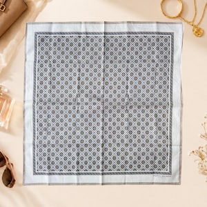 May include: A square Pierre Balmain Paris pocket square featuring a repeating geometric diamond pattern in shades of blue, grey, and white. The fabric has a light blue border and a thin dark grey frame, with the brand name printed in the bottom corner.