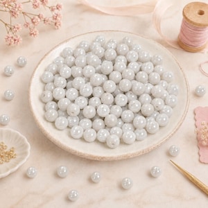 May include: A collection of round, light gray, lustrous beads with a smooth finish. The beads are arranged in a pile on a circular plate, with several individual beads scattered around the plate alongside a spool of pink thread and dried flowers.