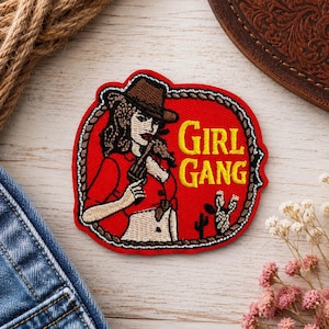May include: Embroidered patch with a red background and a brown border. The patch features a woman in a cowboy hat holding a gun, with the words "GIRL GANG" in yellow. A cactus is also visible.