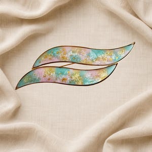 May include: A hair clip featuring two curved, wave-shaped segments with a smooth, glossy finish. The design incorporates a blend of pastel pink and teal colors, accented with embedded gold and silver metallic flakes, all encased in a gold-toned metal frame.