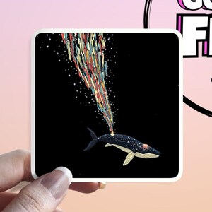 May include: Square sticker featuring a whale with a starry body, spouting a colorful stream of rainbow-colored lines and stars against a black background. The sticker is held by a hand.