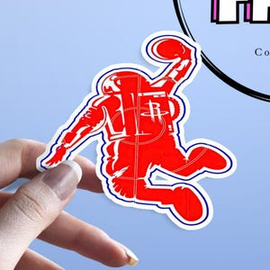May include: A red sticker featuring an astronaut dunking a basketball. The astronaut is wearing a spacesuit and holding a basketball. The sticker has a white border and is held by a hand against a light blue background.