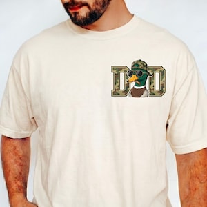 May include: Cream-coloured t-shirt with a graphic of a duck wearing sunglasses and a camouflage hat, and the word "DAD" in camouflage letters. The shirt has short sleeves and a crew neck.