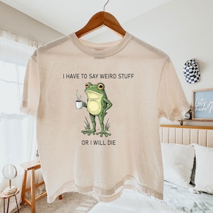 May include: Beige t-shirt with a humorous graphic of a green frog holding a coffee cup. The frog stands in grass, with the text "I HAVE TO SAY WEIRD STUFF OR I WILL DIE" above and below the frog illustration. The shirt is hanging on a wooden hanger.