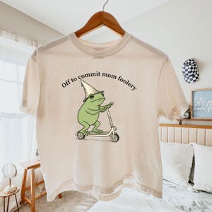 Puede incluir: Camiseta beige con la frase "Off to commit mom foolery" y un dibujo de una rana verde en un patinete blanco y negro, con un sombrero de mago.