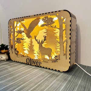 Peut inclure: Veilleuse en bois avec une scène de forêt représentant un cerf, un aigle et des arbres. Le nom "Oliver" est gravé sur la base. La lumière émet une lueur ambrée chaleureuse.