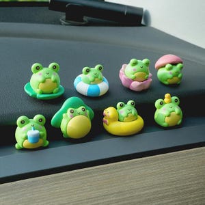 May include: A collection of eight small, green frog figurines with yellow bellies and pink cheeks. The frogs are depicted in various activities, including sitting on a lily pad, floating in a blue and white inner tube, resting in a pink flower, wearing a mushroom cap, holding a blue cup, and balancing a tiny yellow duck on their head.