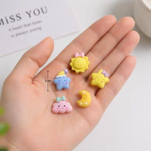 Könnte beinhalten: Fünf kleine, bunte Charms mit flacher Rückseite, bestehend aus einer gelben Sonne mit rosa Schleife, einem blauen Stern mit Schlafmütze, einem gelben Stern mit violetter Schleife, einer rosa Wolke mit grüner Schleife und einem gelben Halbmond, je 2 cm.