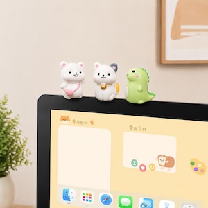 Puede incluir: Tres pequeñas figuras de animales posadas en el borde superior de un monitor de computadora. El conjunto incluye un oso blanco con un bolso rosa, un gato blanco con un collar de campana dorada y un dinosaurio verde claro con acabado liso.