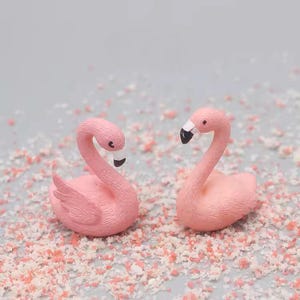Puede incluir: Dos pequeñas figuras de flamencos rosas con plumas texturizadas, cuellos curvados y picos con puntas negras. El flamenco de la izquierda tiene la cabeza ligeramente inclinada hacia abajo, mientras que el de la derecha la mantiene erguida.