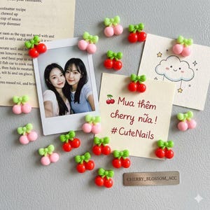 May include: A collection of small cherry-shaped charms, each featuring two red or pink spheres topped with a green bow. These charms are scattered around a photograph of two people and a note with the text Mua thêm cherry nữa #CuteNails.